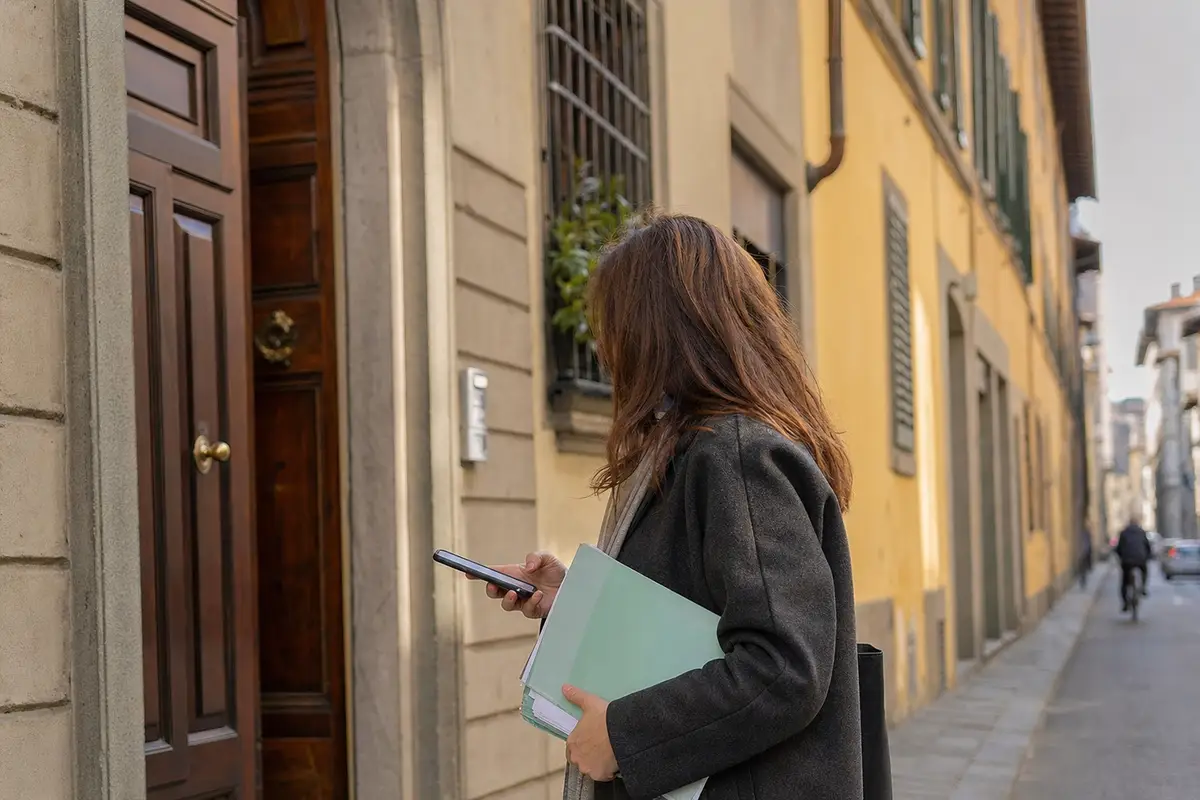 Donna davanti a un portone con smartphone e cartellina di documenti in una strada residenziale a Firenze