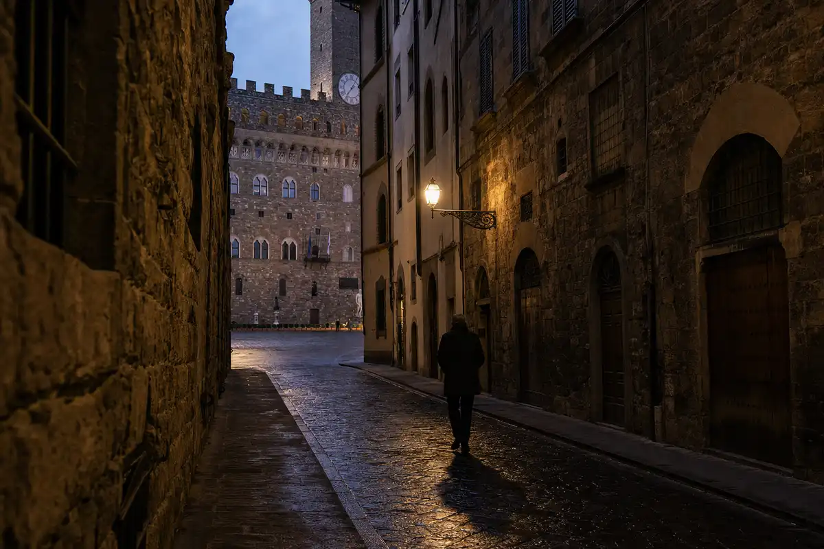Vicolo di Firenze al crepuscolo con pavé bagnato, un passante e Palazzo Vecchio sullo sfondo